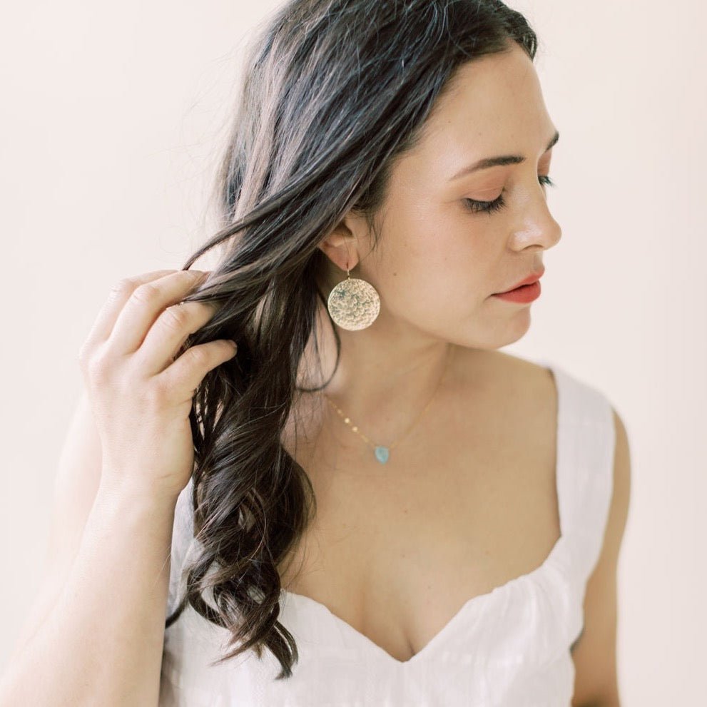 Gold Phoebe Earrings by Sarah Cornwell Jewelry. Dark haired woman wearing white top and shimmery, textured 1.25 inch gold disc statement earrings and chalcedony pendant necklace .