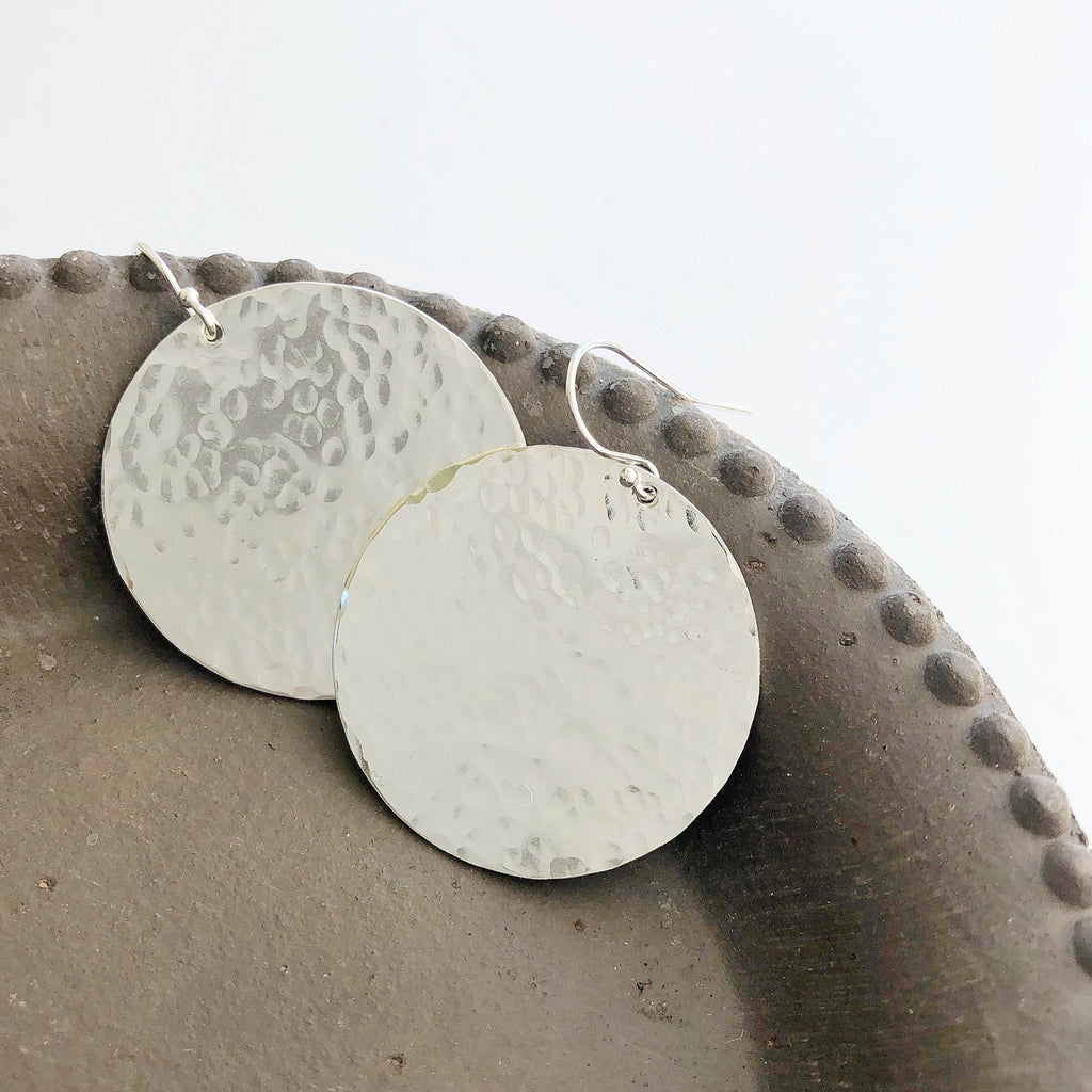 Silver Phoebe Earrings by Sarah Cornwell Jewelry. Shimmery, textured 1.25 inch silver disc statement earrings shown closeup on a gray plate.
