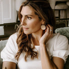 Gold Phoebe Earrings by Sarah Cornwell Jewelry. Blonde haired woman sitting on sofa wearing white short sleeved top and shimmery, textured 1.25 inch gold disc statement earrings and gold chain necklace .