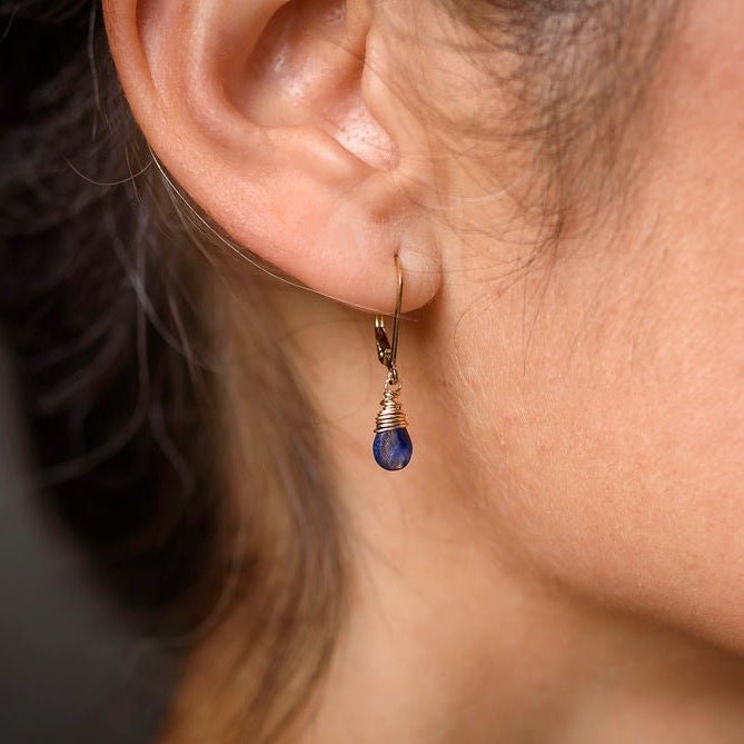 Poppy Blue Earrings by Sarah Cornwell Jewelry. Side view of a woman's ear wearing gold wire wrapped blue sapphire lever back .75 inch drop earrings.