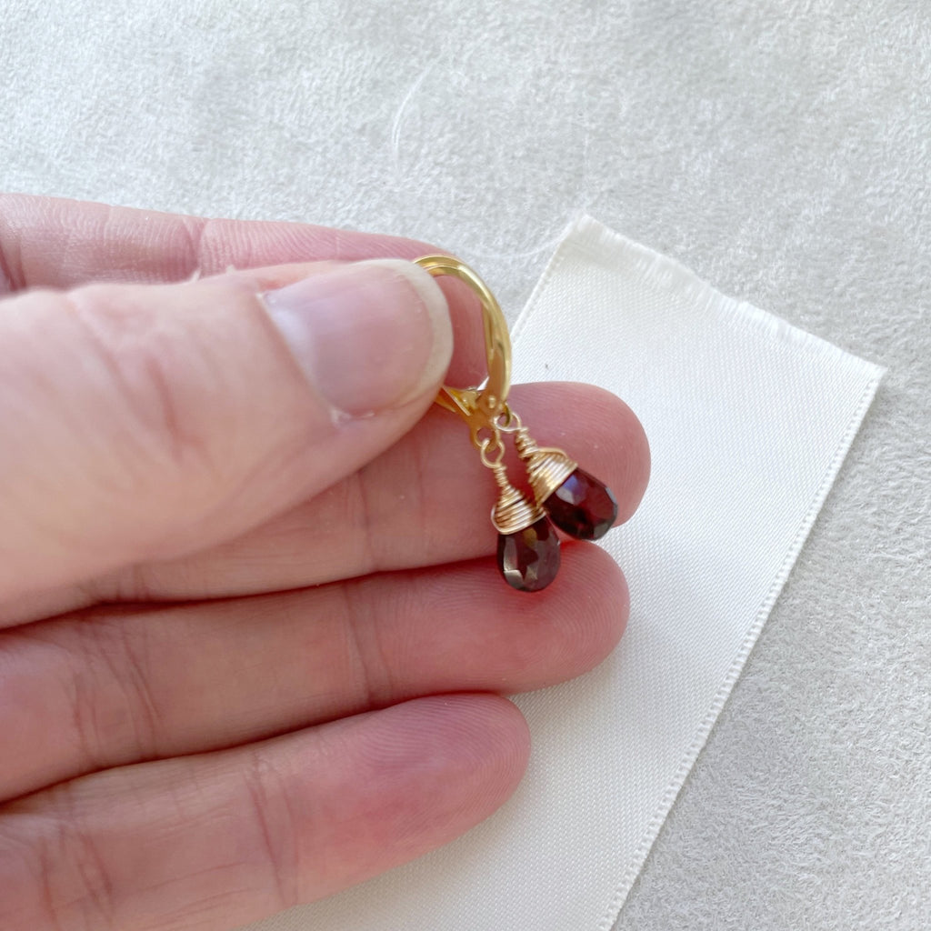 Hand holding gold wire wrapped garnet gemstone drop earrings with a lever back. Poppy Garnet Earrings by Sarah Cornwell Jewelry