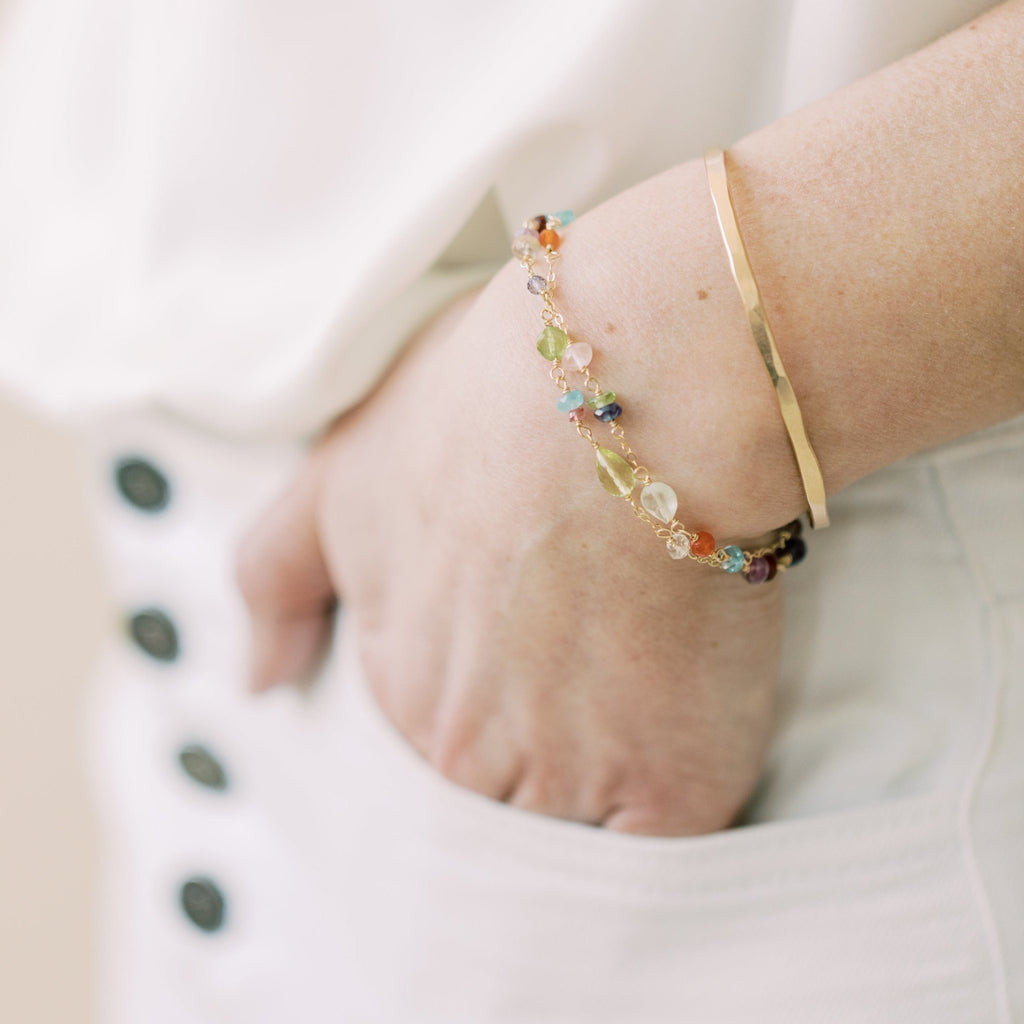 Close up of woman's wrist wearing white pants and white shirt with gold and silver rainbow gemstone bracelet with wire wrapped aquamarine, peridot, quartz, amethyst, sapphires, chalcedony, citrine, smokey quartz, garnet, and labradorite gemstones and gold bangle. Poppy Love Bracelet by Sarah Cornwell Jewelry