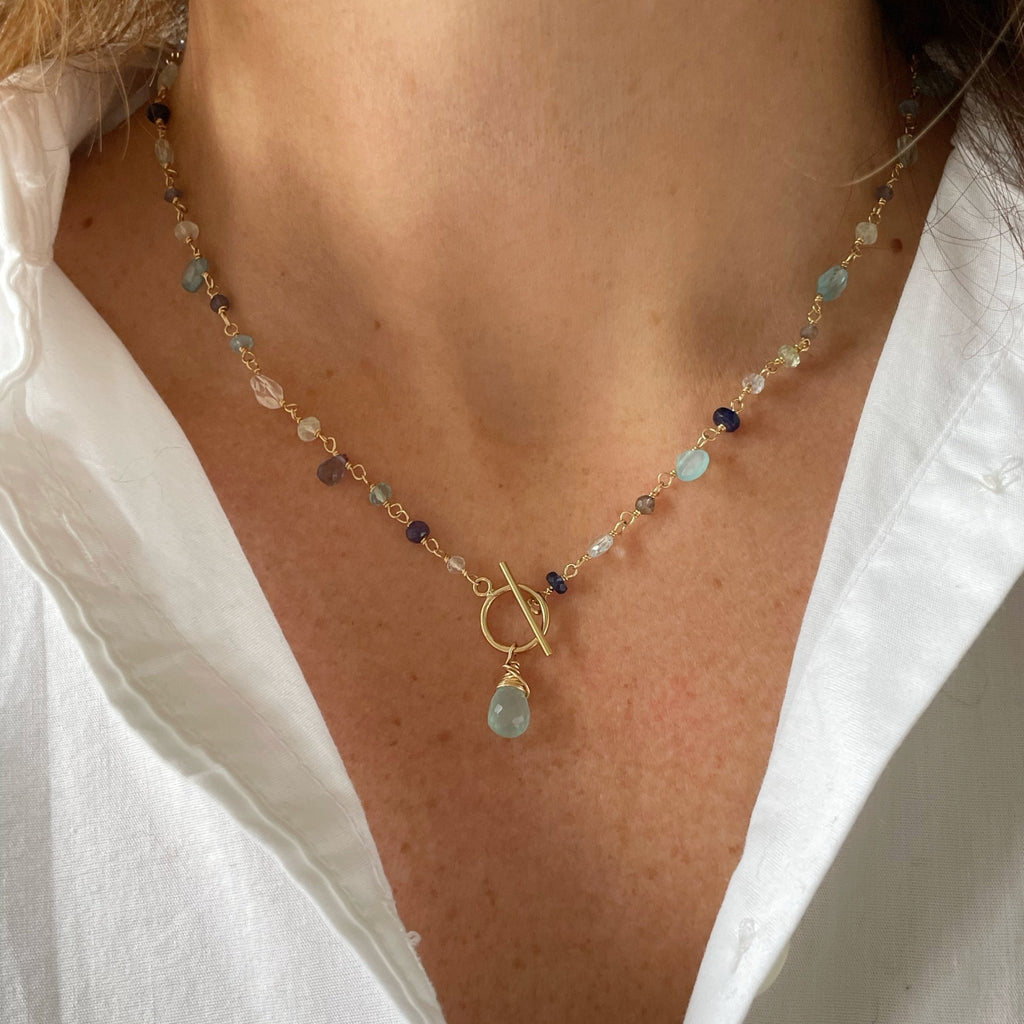 Women's neckline wearing white button down with gold gemstone necklace with wire wrapped blue chalcedony, aqua chalcedony, prehnite, iolite, blue sapphires, and aquamarine gemstones with a front toggle closure and large chalcedony drop. Poppy Seaside Front Toggle by Sarah Cornwell Jewelry