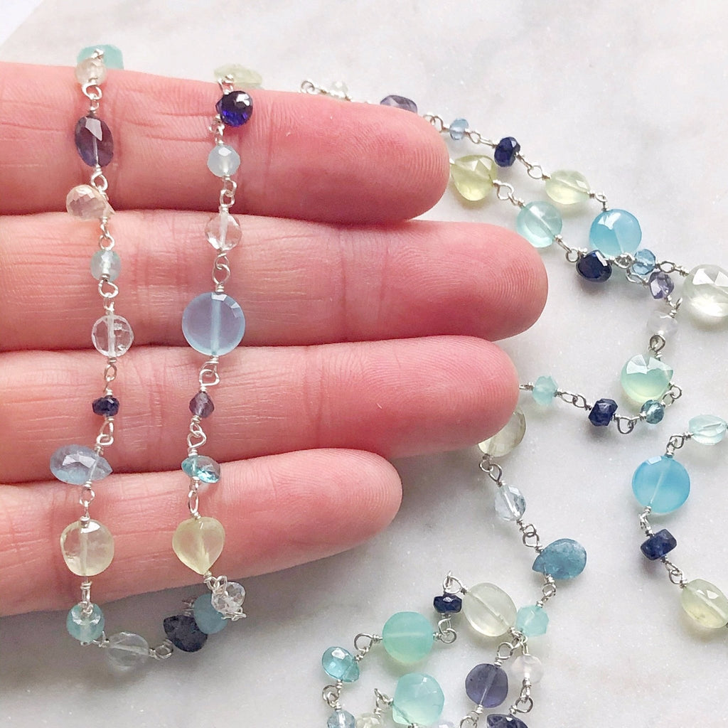 Hand holding silver and gemstone long statement necklace with wire wrapped blue chalcedony, aqua chalcedony, prehnite, iolite, blue sapphires, and aquamarine gemstones. Poppy Seaside Necklace by Sarah Cornwell Jewelry