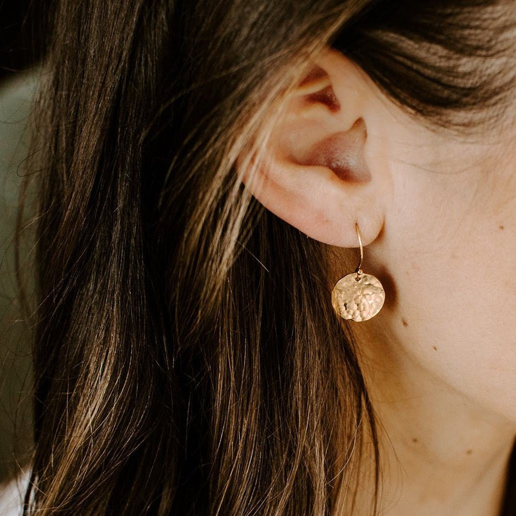 Gold Rachel Earrings by Sarah Cornwell Jewelry. Side view of dark haired woman's ear wearing shimmery, dainty gold 1/2 inch textured disc earrings.