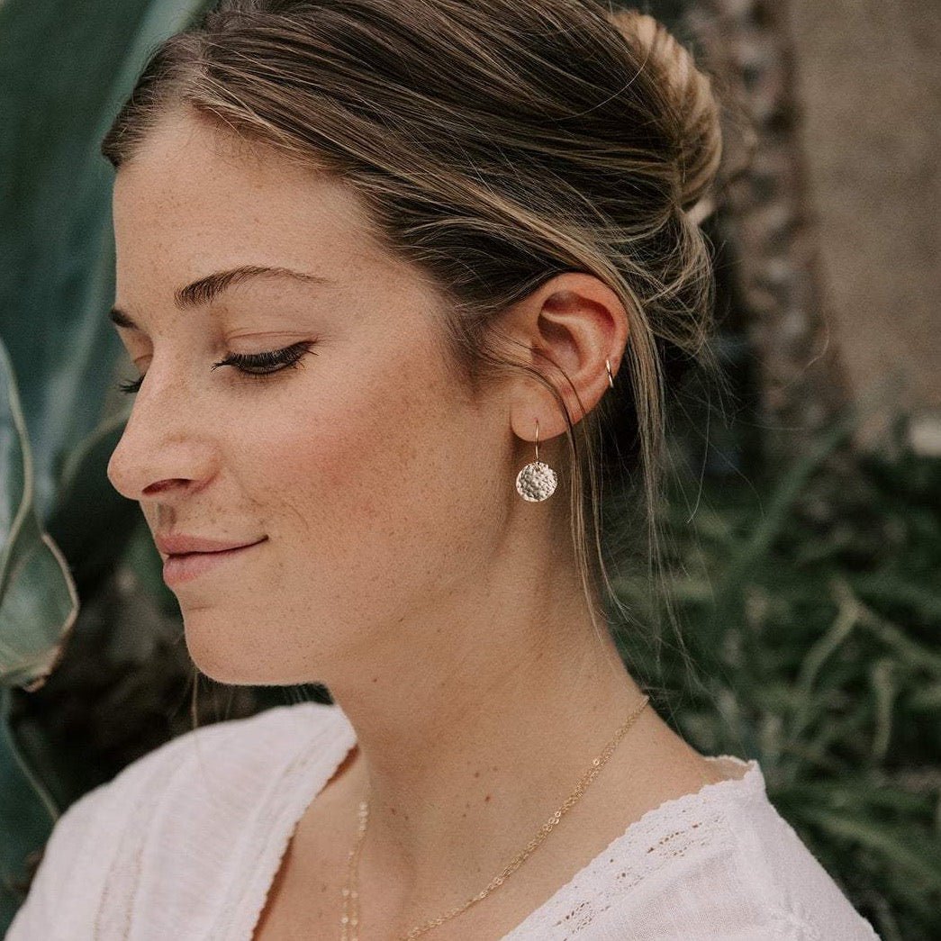Gold Rachel Earrings by Sarah Cornwell Jewelry. Side view of blonde woman wearing a white top and shimmery, dainty gold 1/2 inch textured disc earrings and gold chain necklace.