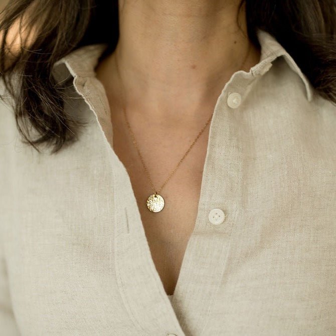 Woman's neckline wearing beige linen button down shirt with gold textured disc necklace. Ross Necklace by Sarah Cornwell Jewelry