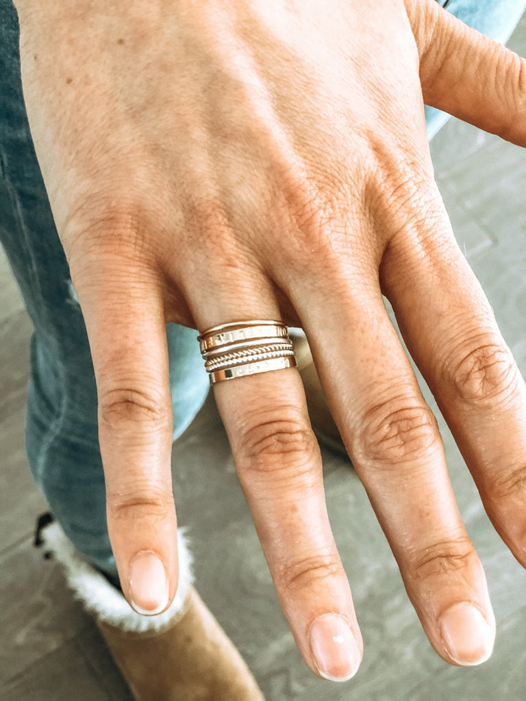 Close up of woman's hand wearing 6 gold rings of various styles. One ring has capitol letter names stamped around it. Rowan Ring by Sarah Cornwell Jewelry