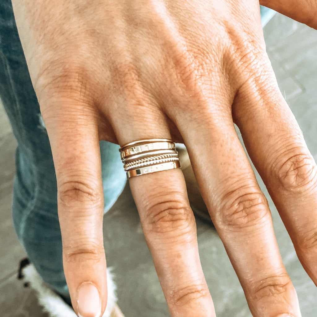 Close up of woman's hand wearing 5 gold rings of various styles. One ring has capitol letter names stamped around it. Ruby Stacked Rings by Sarah Cornwell Jewelry