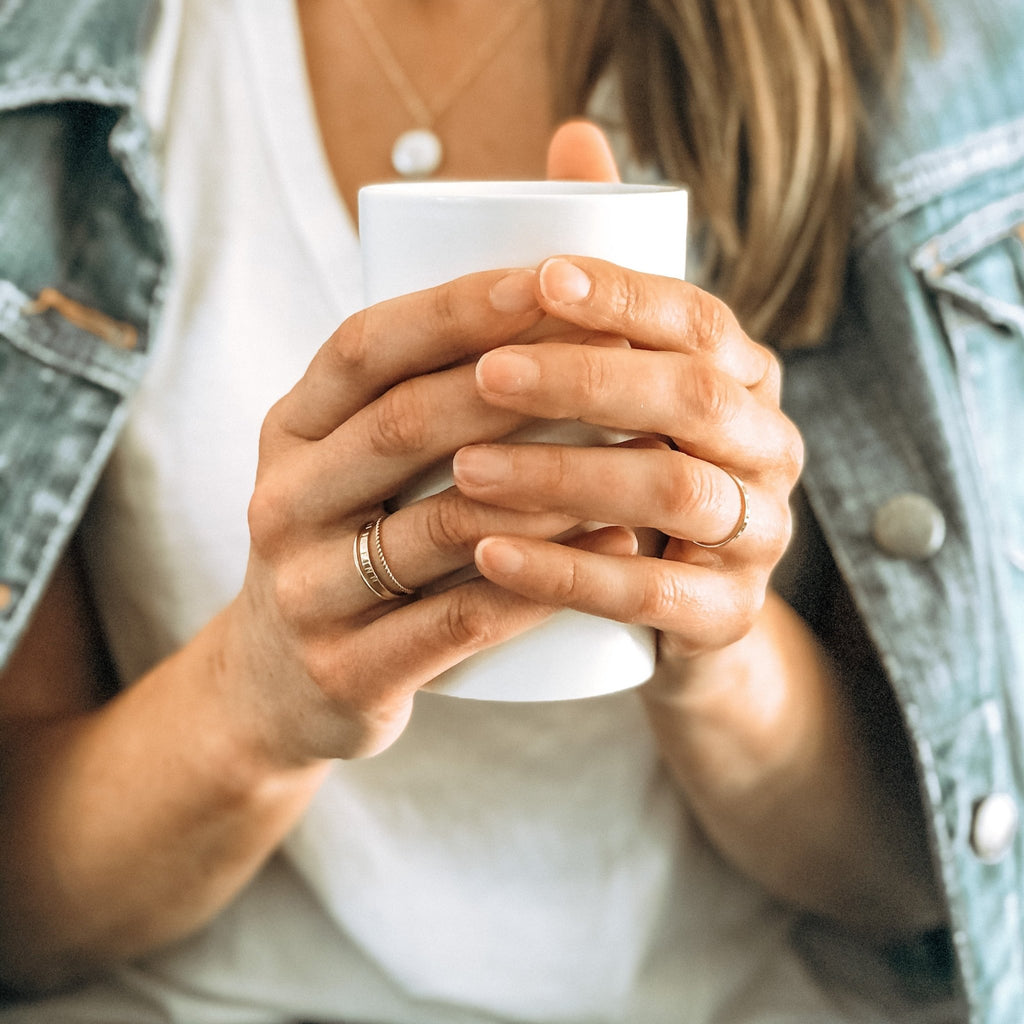 Close up of woman's hands holding a cup of coffee wearing 3 gold rings of various styles. One ring has capitol letter names stamped around it and one single ring on the other hand with a gold coin pearl necklace and denim jacket. Ruby Stacked Rings by Sarah Cornwell Jewelry