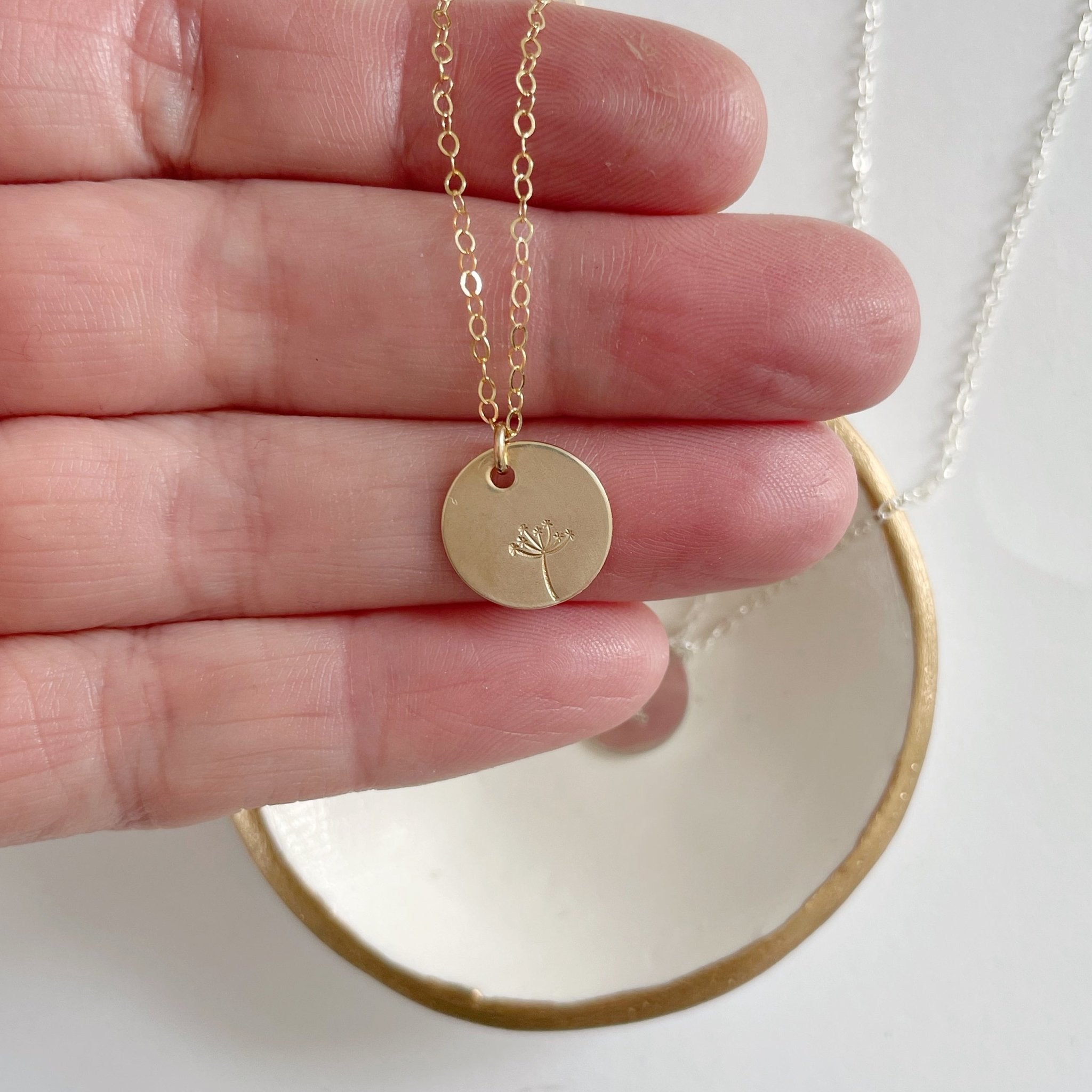 Hand holding gold stamped disc dandelion seed miscarriage necklace. Seeds of Hope Necklace by Sarah Cornwell Jewelry