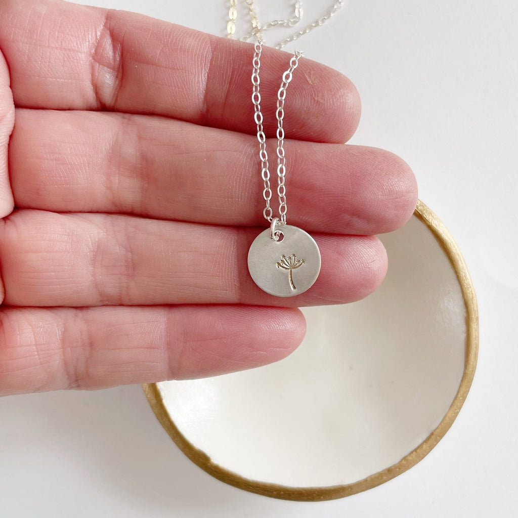 Hand holding silver stamped disc dandelion seed miscarriage necklace. Seeds of Hope Necklace by Sarah Cornwell Jewelry