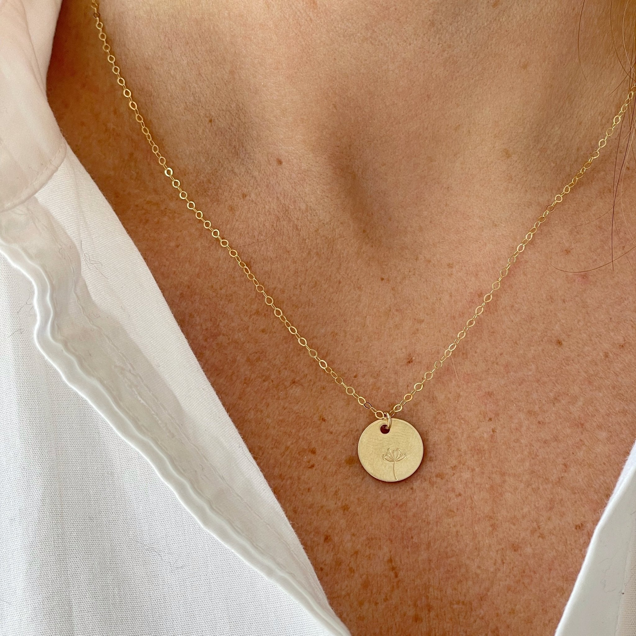 Close up of woman's neckline wearing white shirt with gold stamped disc dandelion seed miscarriage necklace. Seeds of Hope Necklace by Sarah Cornwell Jewelry