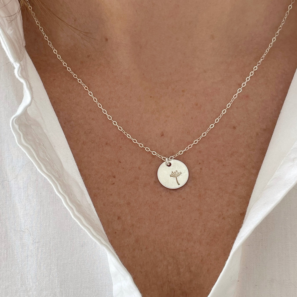 Close up of woman's neckline wearing white shirt with silver stamped disc dandelion seed miscarriage necklace. Seeds of Hope Necklace by Sarah Cornwell Jewelry