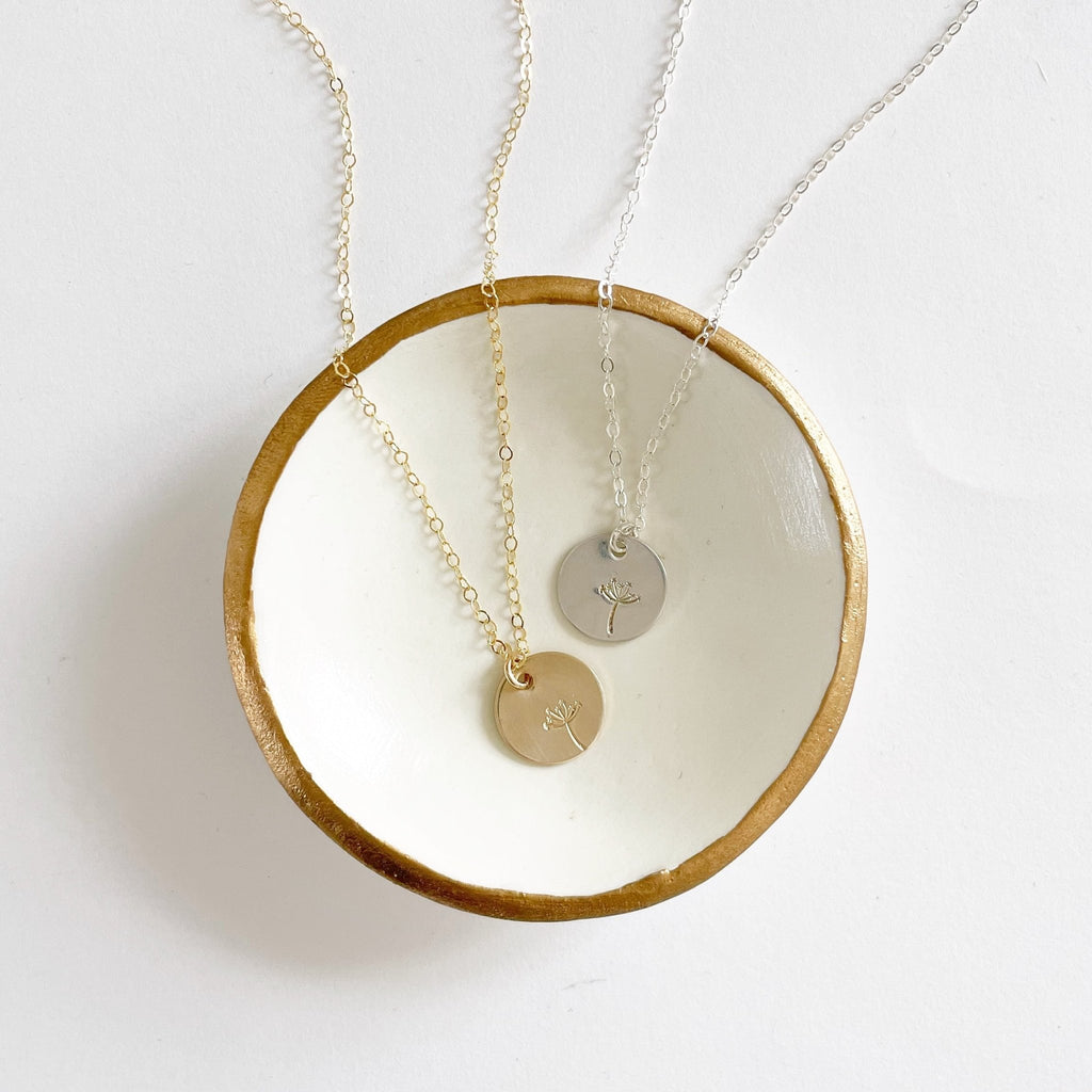 2 Gold and silver stamped disc dandelion seed miscarriage necklaces in a white and gold bowl. Seeds of Hope Necklace by Sarah Cornwell Jewelry