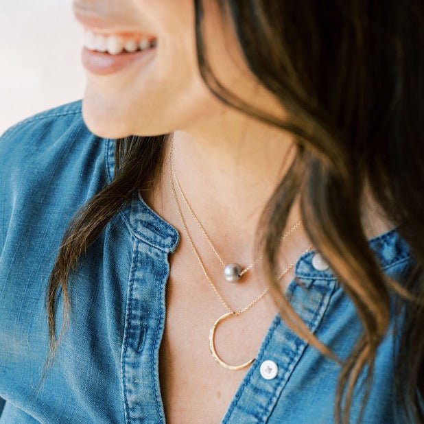 Close up of woman's neckline wearing denim top with gold chain necklace with single large silver gray freshwater pearl and moon shaped gold pendant necklace. Tomlin Necklace by Sarah Cornwell Jewelry