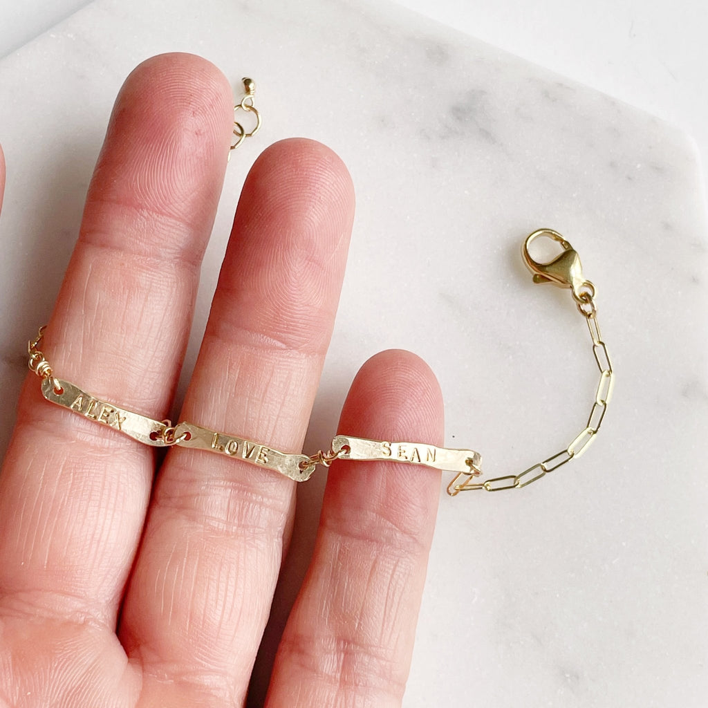 Hand holding gold link chain bracelet with 3 hand forged bars with hand stamped names. Tribe Bracelet by Sarah Cornwell Jewelry