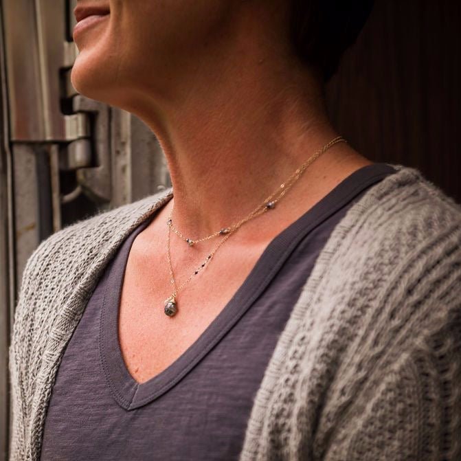 Woman's neckline wearing gray shirt and taupe sweater with gold sapphire, gray zircon and tiny pearl gemstone layering necklace and gold gemstone layering necklace. Walden Necklace by Sarah Cornwell Jewelry